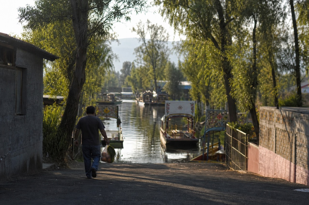 A traveler walking down a paved lane toward a canal dock entrance carrying a bag of groceries