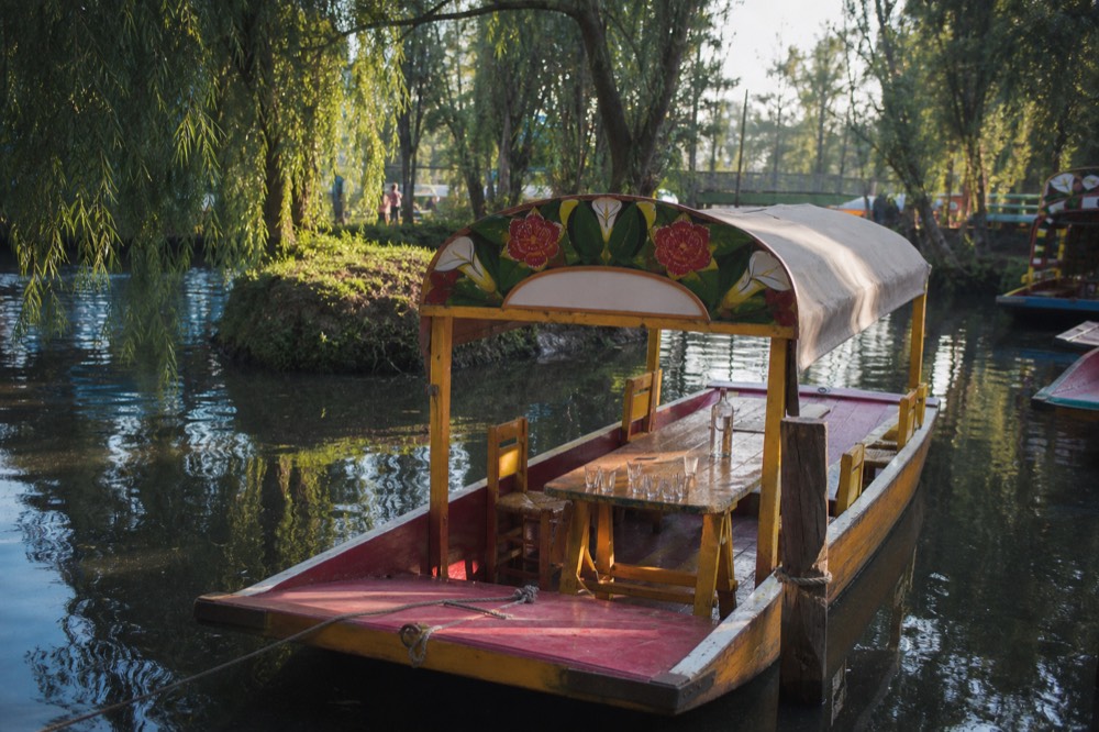 A canopied canal boat tied to a wooden post beside an overgrown island bank, shot glasses and a bottle on the long table inside