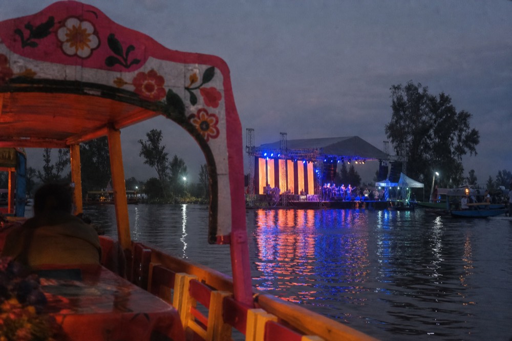 A floating stage lit in blue and orange seen across dark water from the rail of a boat at night