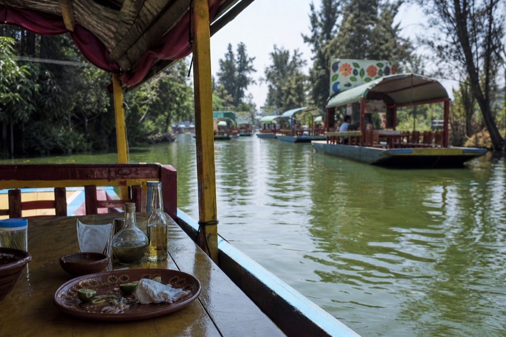 A wooden plank table inside a canal boat set with plates, cut limes and bottles as the green water slides past outside