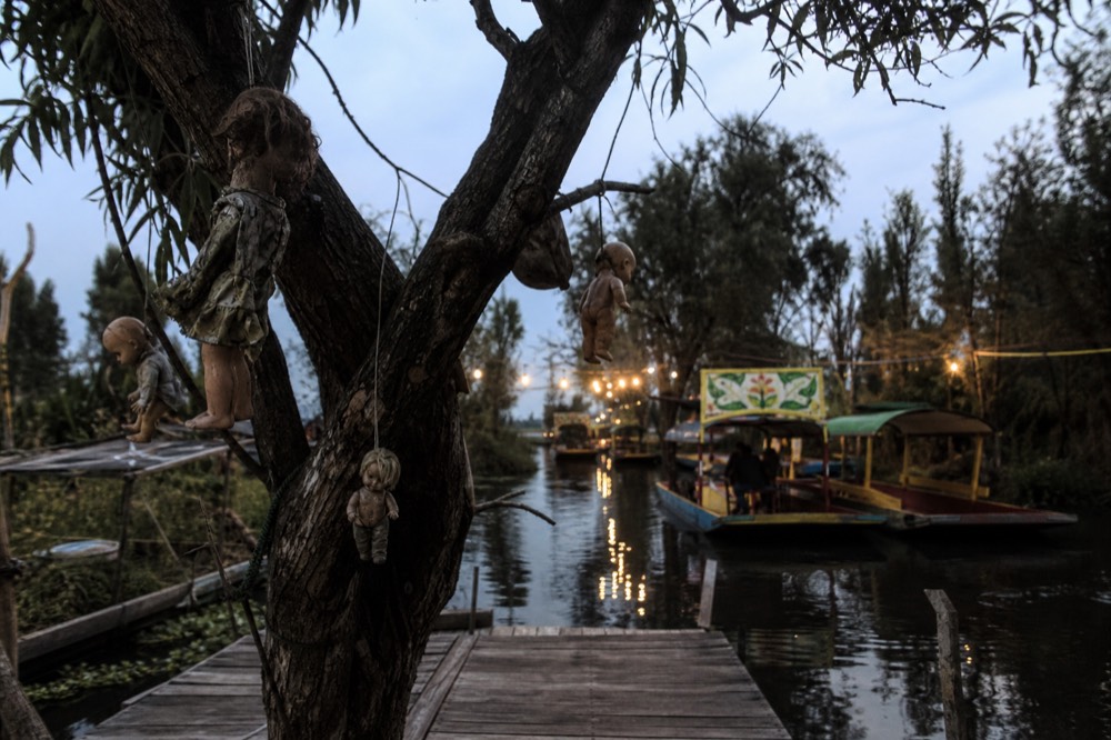 Weathered dolls tied to tree branches above a wooden platform at the edge of a narrow overgrown canal