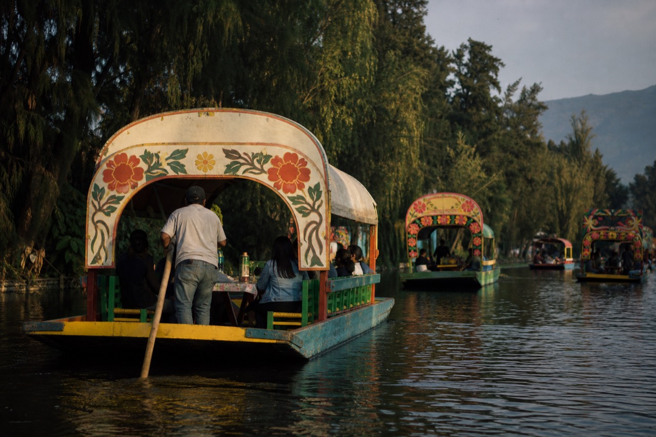 A long canopied canal boat seen from behind as it moves down an open stretch of water lined with willow trees