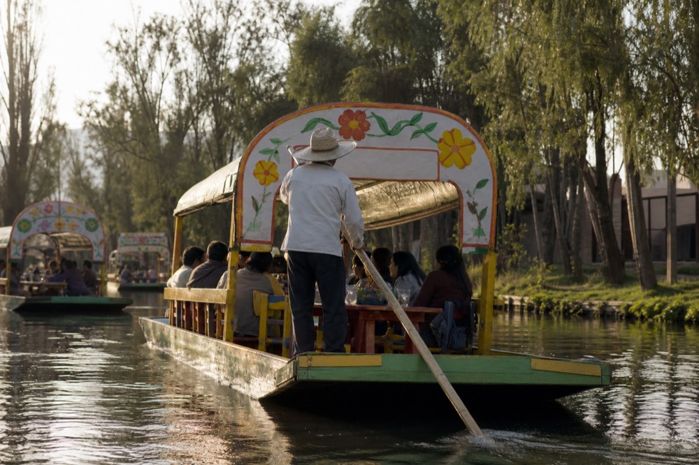 A poler in a straw hat pushing a long wooden pole against the canal bed while passengers sit at a long table beneath the boat canopy