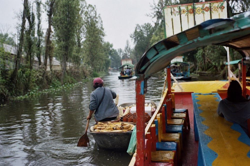 A small paddled canoe loaded with grilled corn cobs and clay cups pulling alongside a larger canopied boat on the water