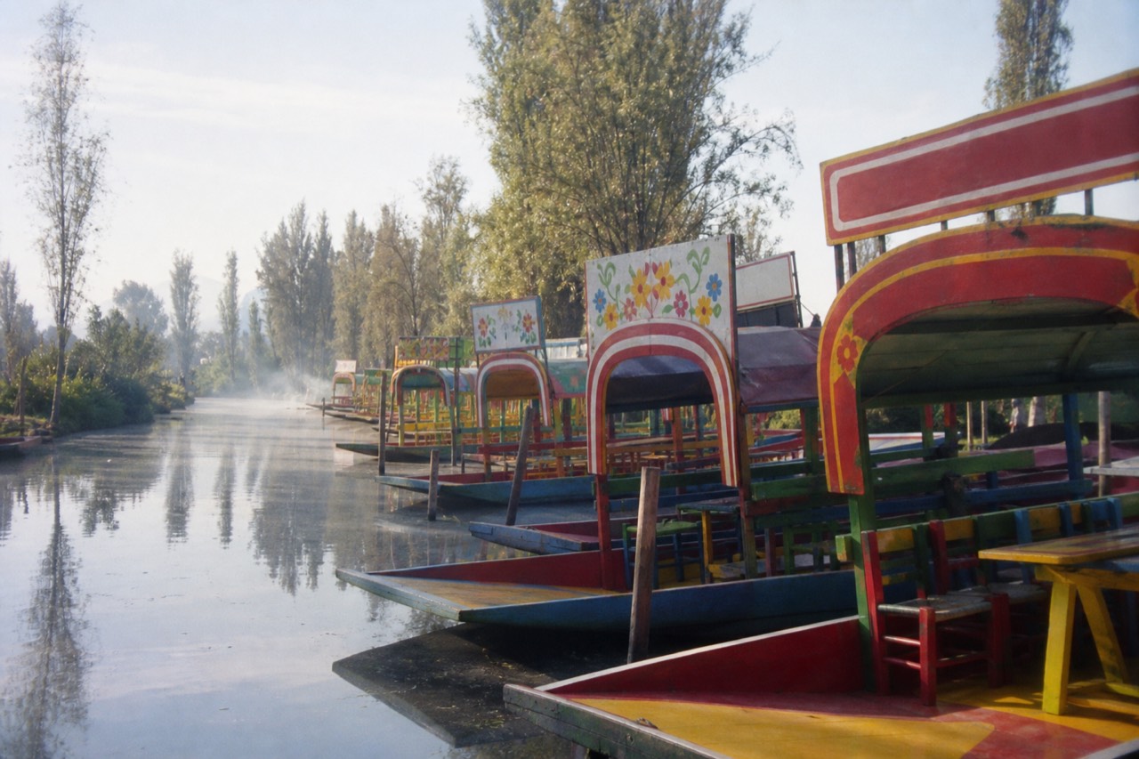 Rows of brightly painted flat-bottomed boats with arched wooden canopies tied along a canal dock under a bright afternoon sky