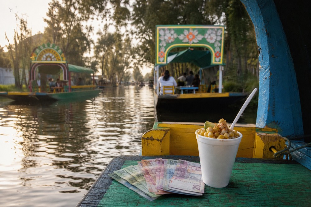 Fanned Mexican peso banknotes in small denominations resting on a painted wooden boat bench beside a paper cup of esquites