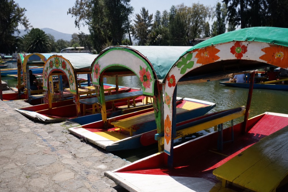 Rows of brightly painted flat-bottomed canal boats with arched canopies moored along a stone landing under midday sun