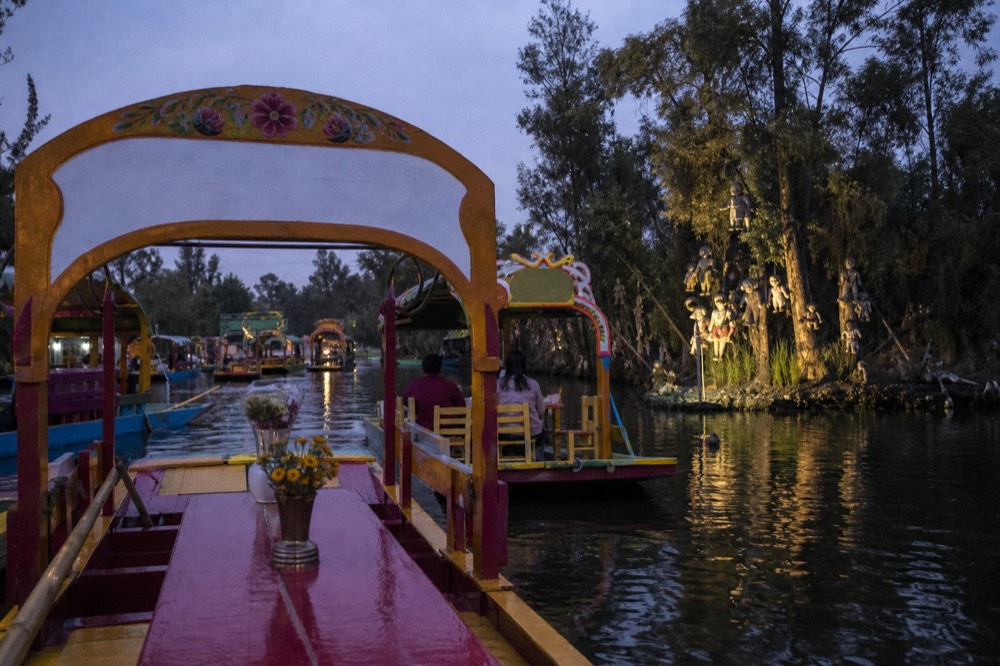 Film-style night scene of a canal island lit from below, dolls in the trees casting long shadows across dark water