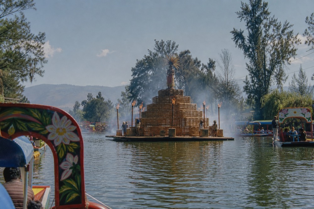 A pyramid-shaped wooden stage rising from open water at night, ringed by burning torches and drifting mist