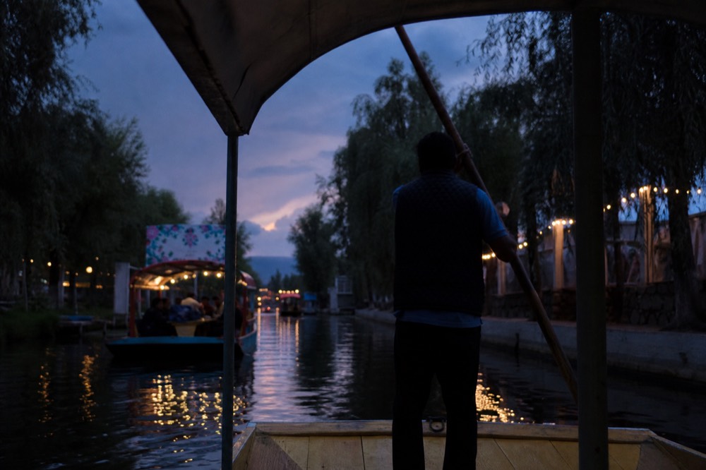 A boatman in silhouette leaning on a long wooden pole, guiding a canopied boat down a narrow canal lined with black willows at dusk
