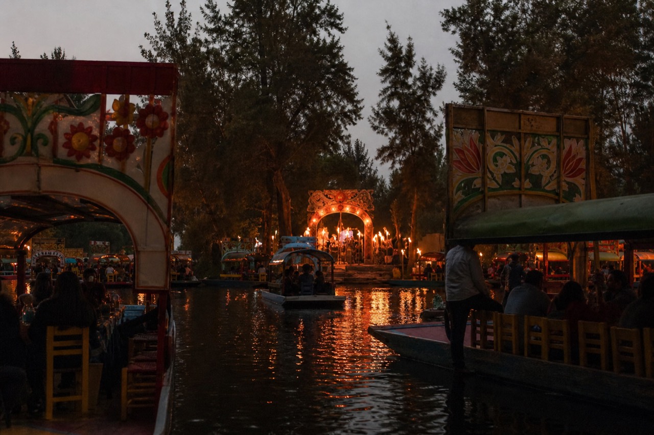 Canal boats gathered in darkness around a lit floating stage on a lagoon, torch flames reflecting on black water