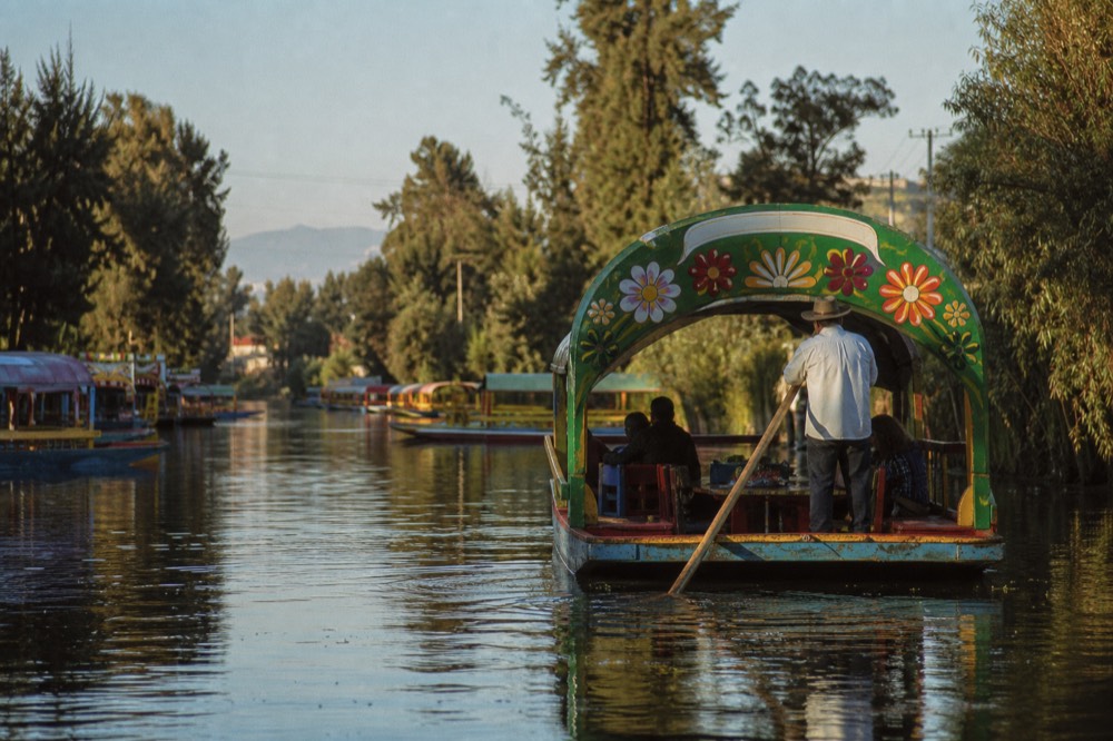 Late afternoon canal in warm golden light, a roofed wooden boat silhouetted against water with a musician standing at the bow
