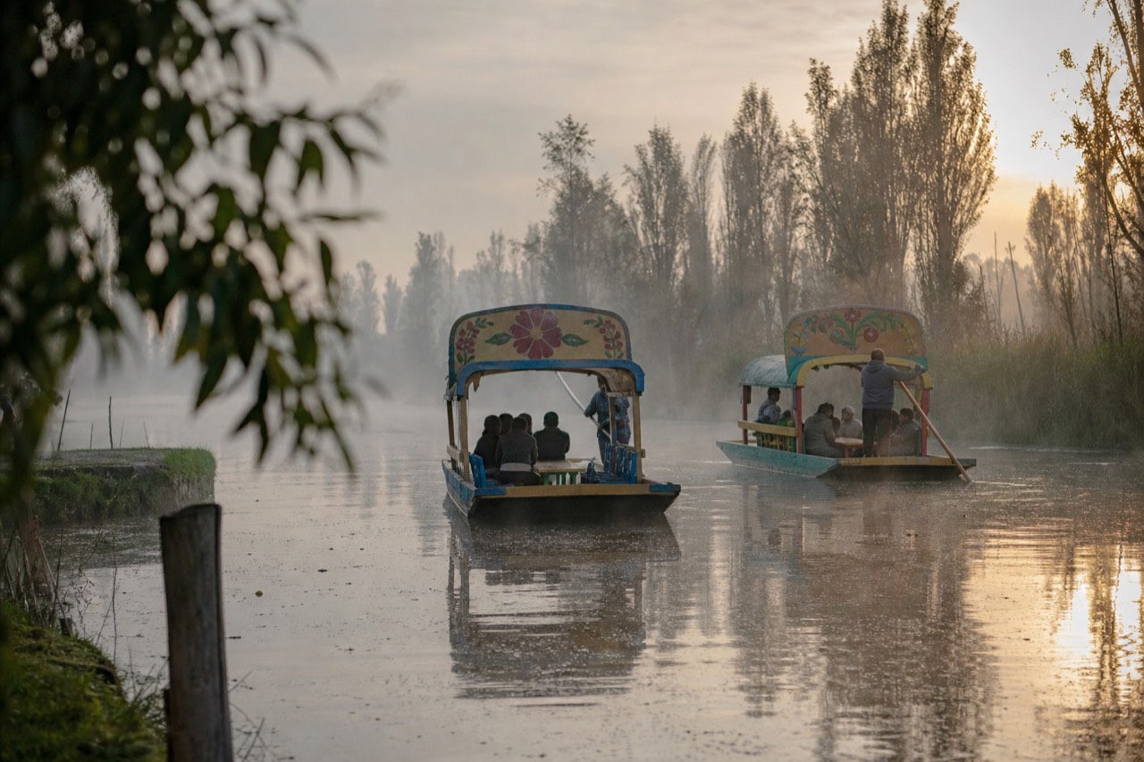Two brightly painted flat-bottomed canal boats passing in early morning mist, low sun behind reed-lined banks