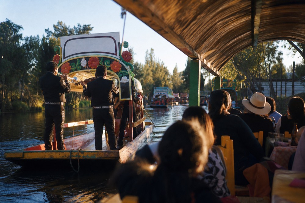 Musicians in matching charro suits playing trumpet and violin from a small boat pulled alongside a larger roofed canal boat full of seated guests