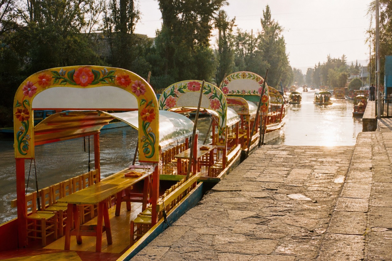 Row of vividly painted canal boats with flower-decorated arched roofs moored along a stone dock under a bright sky
