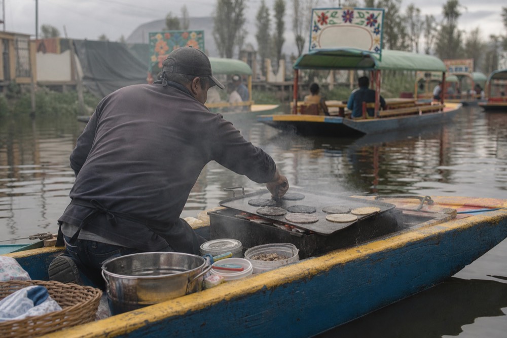 A cook kneeling in a narrow canoe turning blue-corn tortillas on a flat metal griddle over coals, steam rising over the water