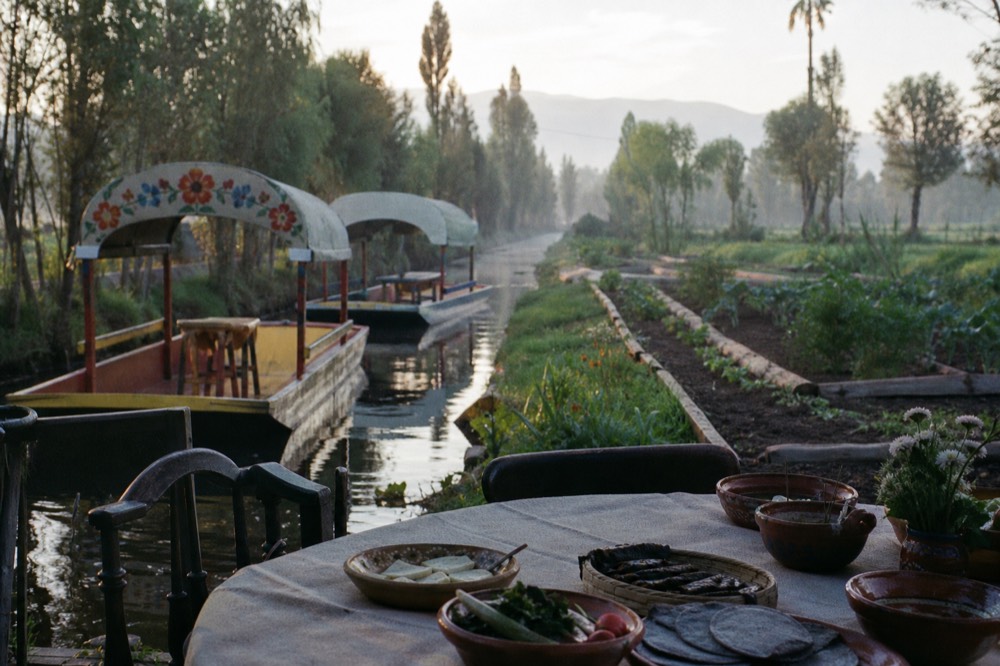 A long table set with clay bowls and blue-corn tortillas on a farm island, raised vegetable beds and canal water directly behind it