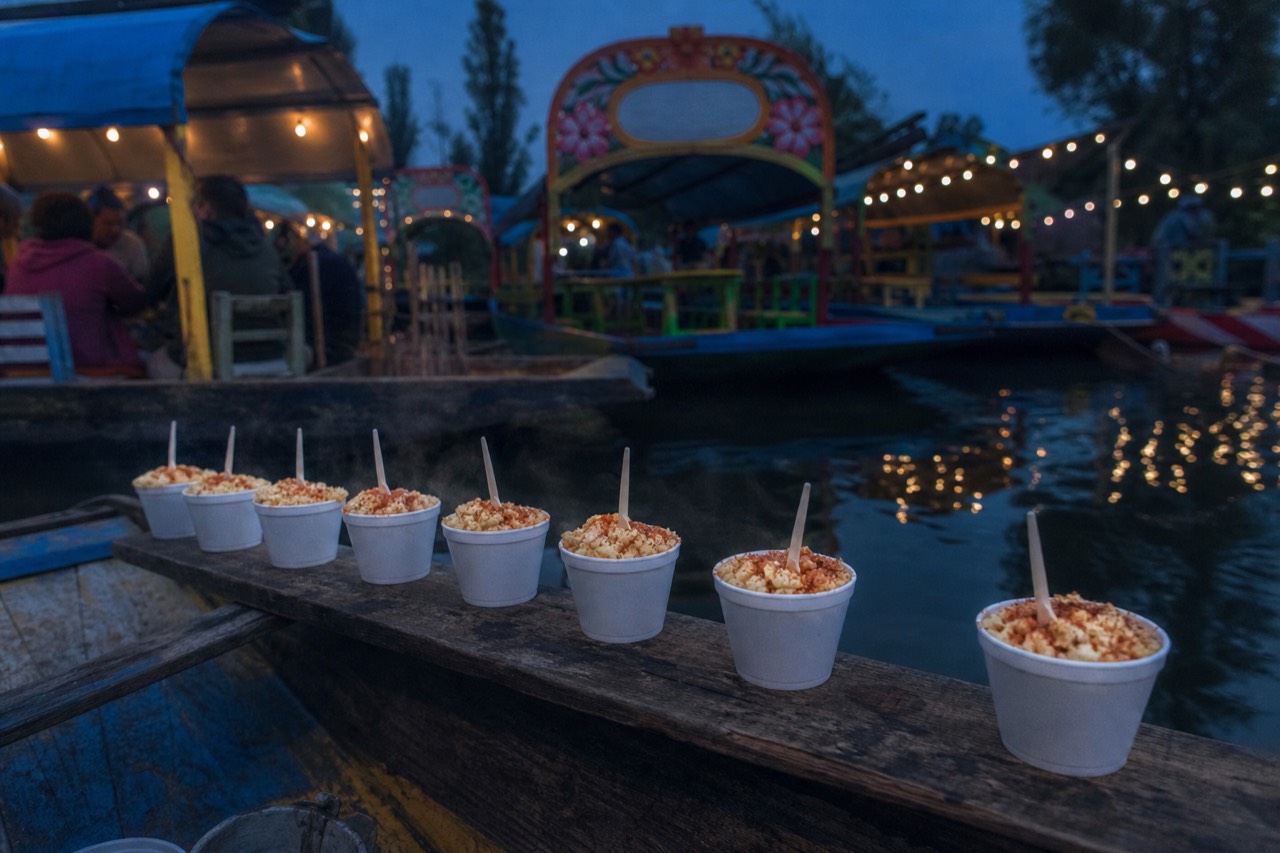Steaming cups of corn kernels topped with cheese and chilli powder lined up on the plank of a small paddled canoe
