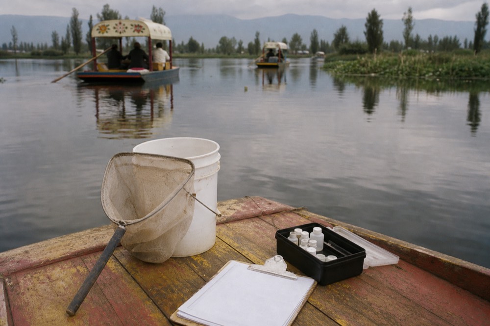 Researcher's field net and sampling bucket resting on the wooden deck of a small boat beside a clipboard and water testing kit