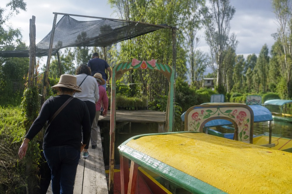 A small group stepping off a canal boat onto a plank landing at a farm island, shade canopy and hand-painted wooden gate ahead