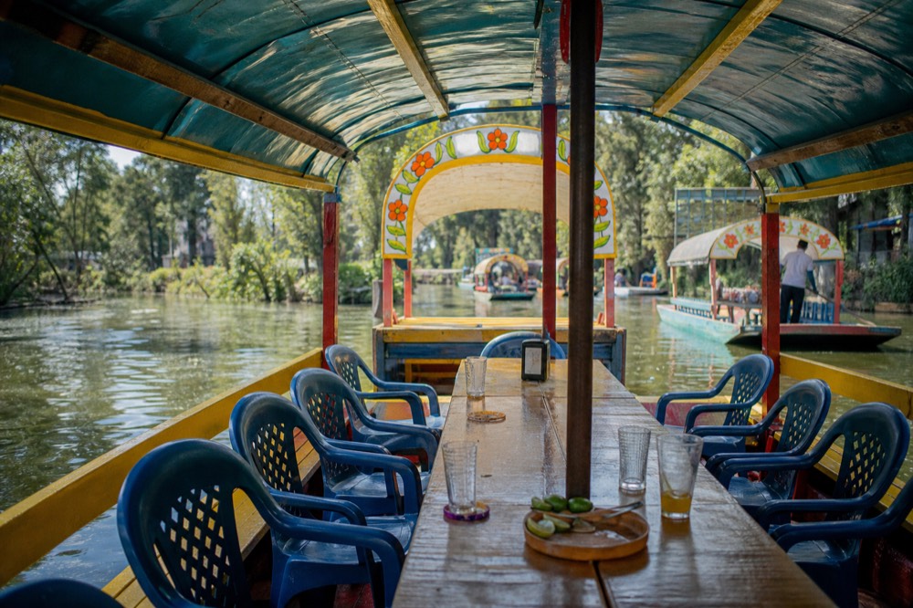 Long wooden table running down the centre of a canal boat under a painted canopy, plastic chairs on both sides, drinks and cut limes on the table
