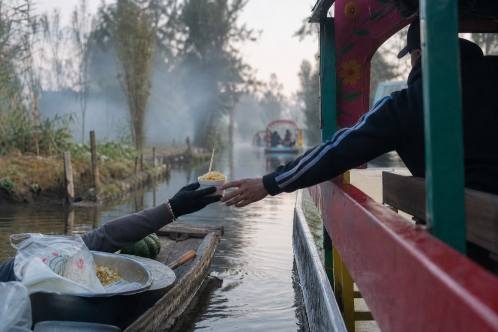 Small paddled canoe pulled alongside a larger canopied boat, a vendor's hands passing across a foam cup of corn kernels topped with cheese