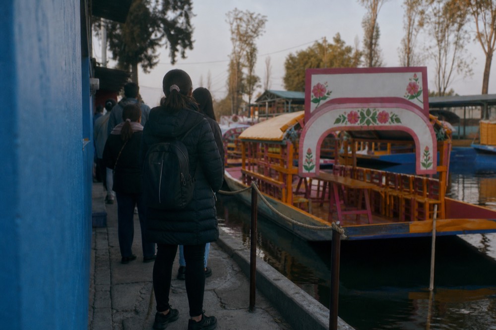 Small group of visitors waiting quietly on a narrow pavement beside a bright blue exterior wall, morning light and a queue barrier rope