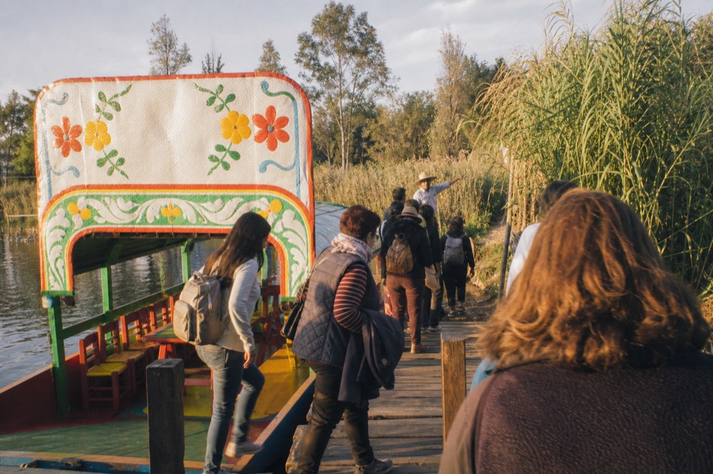 Small tour group of about a dozen people stepping off a canal boat onto a wooden landing, guide gesturing toward a path between reed beds