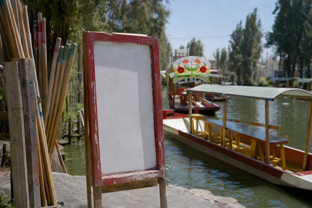 Hand-lettered price board at a canal landing beside a rack of wooden poles, with a moored boat and green water behind