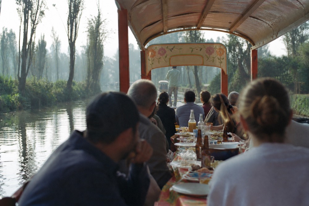 Family group settled around a long table on a moving canal boat, plates and bottles between them, water and green banks sliding past