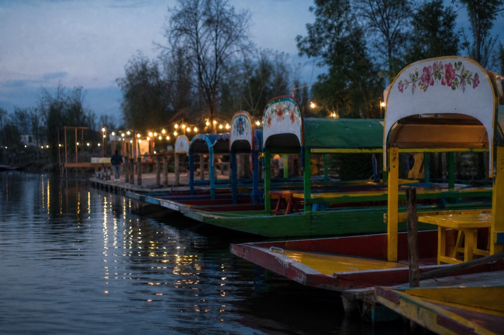 Boats moored bow-in at dusk along a landing with lamps just lit, empty chairs on the decks and reflections stretching across still water