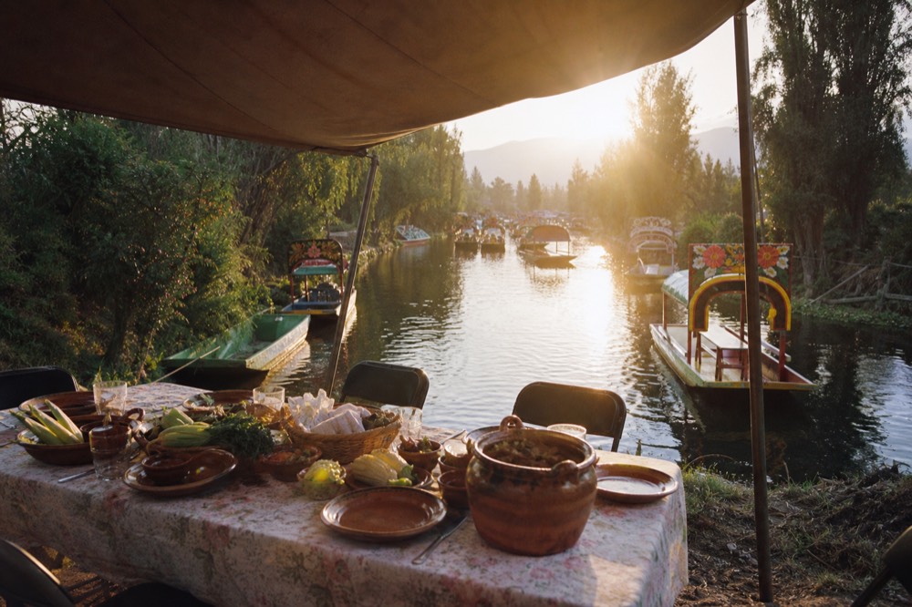 A long table set for lunch under a canvas shade on a farm island, clay dishes and fresh vegetables laid out, canal behind