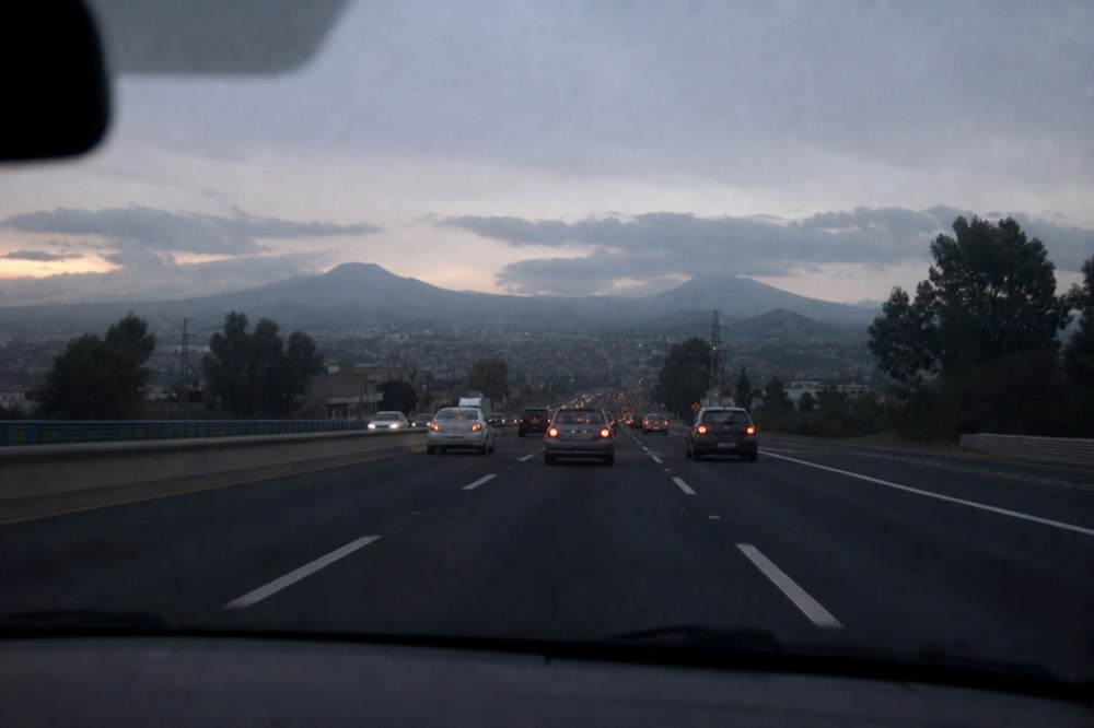View through a car windscreen of a wide southbound highway in Mexico City at dawn, brake lights ahead and volcanic hills on the horizon