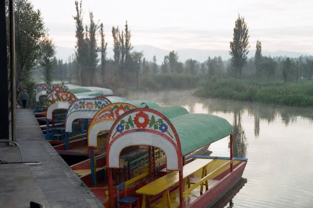 Row of brightly painted wooden trajinera boats moored along a concrete dock, their arched canopies lettered with women's names in flowers