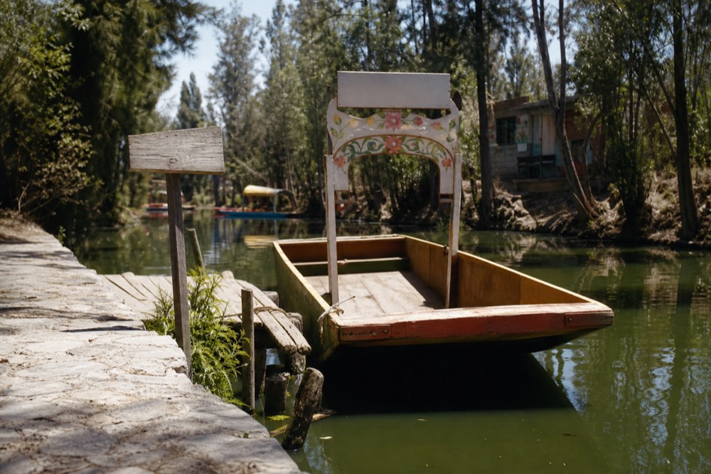 A stone-edged canal bank with a hand-lettered wooden sign post, small wooden landing stage and a farmer's flat cargo boat tied up