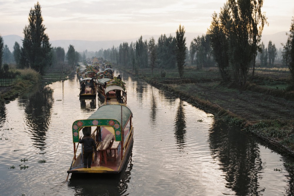 A junction where three canals meet, one crowded with brightly painted passenger boats and the other two empty and lined with farm plots