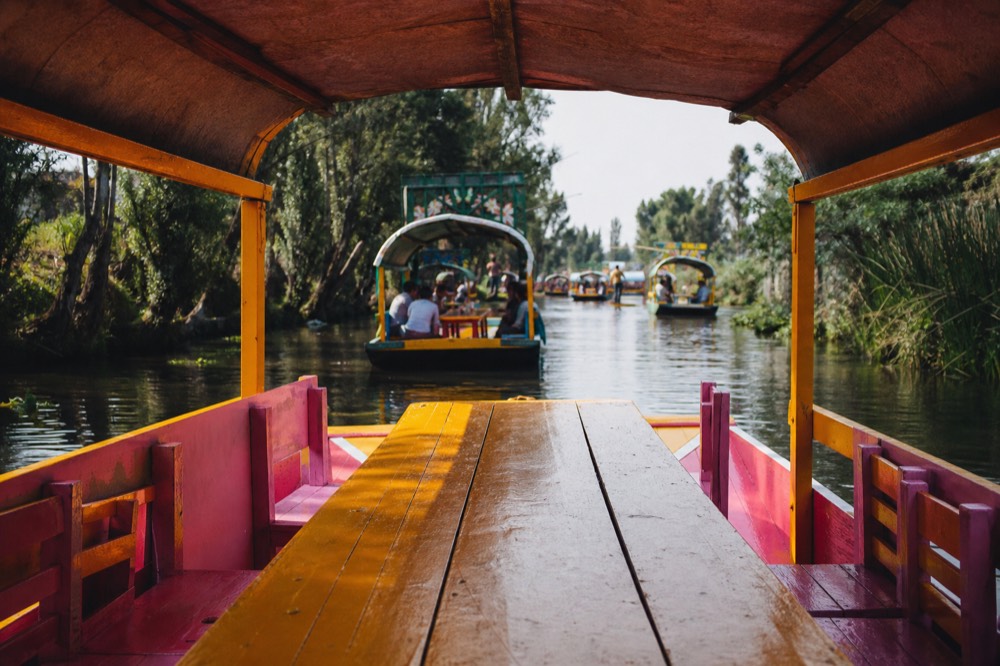 View down the length of a canal boat's long table toward the open bow, other boats ahead and green banks either side under midday sun