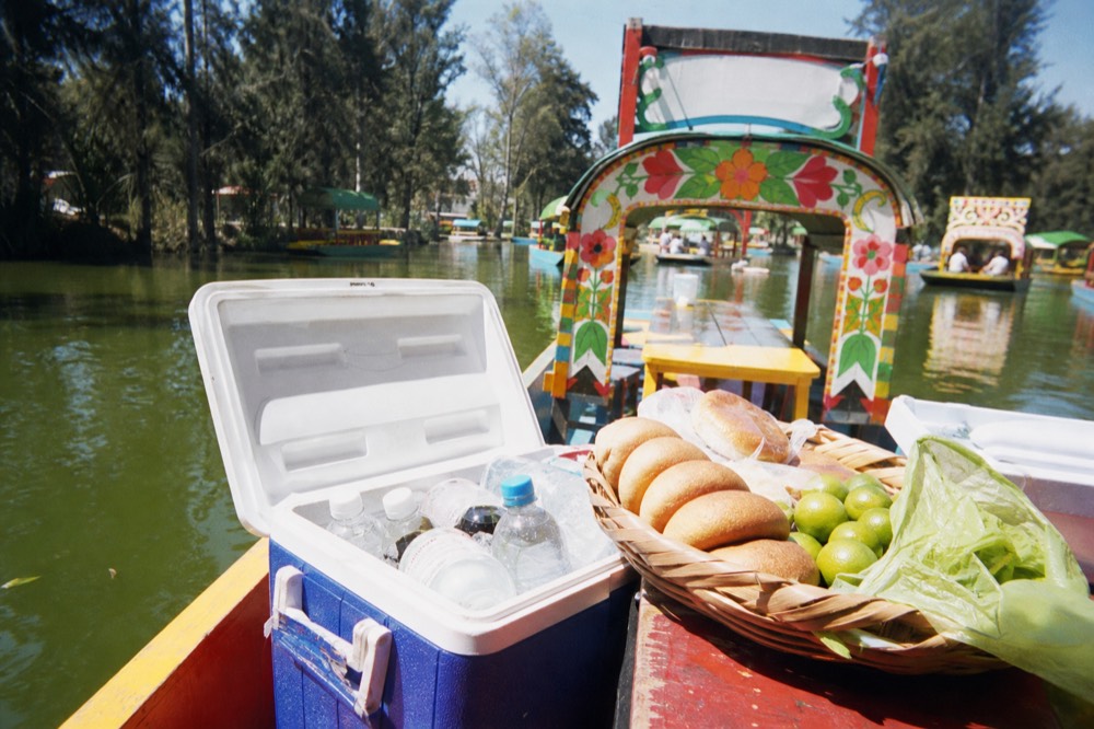 Open cooler packed with ice and bottles beside a woven basket of bread rolls and limes on a boat's wooden bench