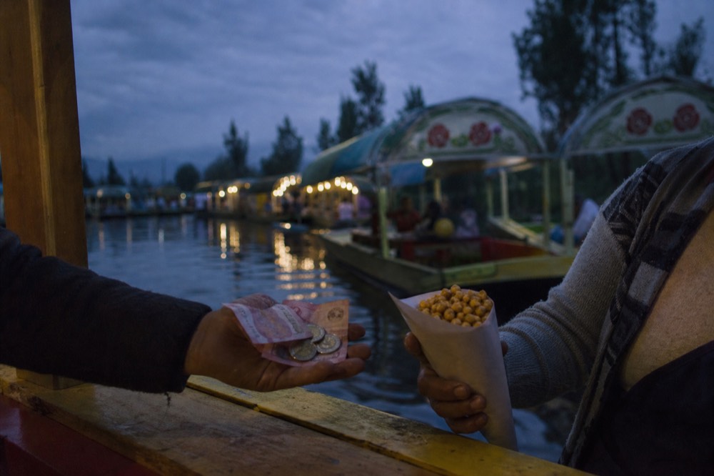 Close view of peso notes and coins being handed across a boat rail to a vendor holding a paper cone of roasted corn