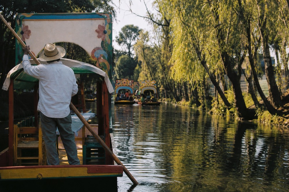 Boatman seen from behind at the stern of a flat-bottomed canal boat, pushing off with a long wooden pole, ripples spreading across dark green water