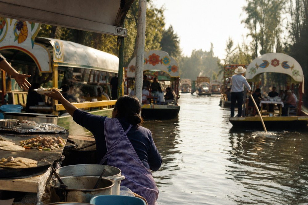 A floating kitchen canoe with a hot griddle and steaming pots pulled tight against a canopy post, a plate being passed up over the gunwale