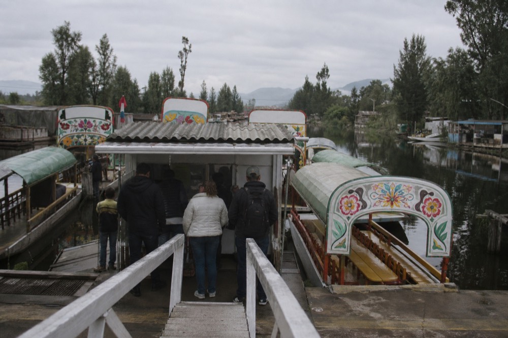 Ticket kiosk at the head of a canal dock seen from the entrance ramp, visitors waiting with their backs to the camera and canopied boats moored bow-in behind