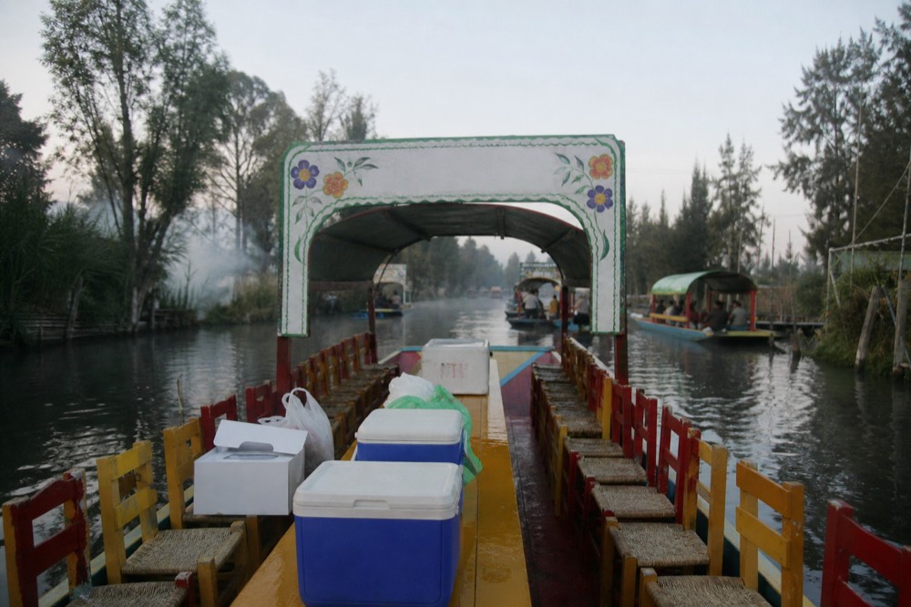 Cooler boxes, a white cake box and shopping bags stacked at the end of a boat's long table before departure, chairs set out along both sides