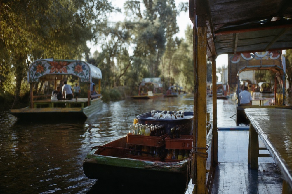 A vendor canoe loaded with crates of bottled drinks and a tub of ice, tied briefly against a larger boat's canopy post