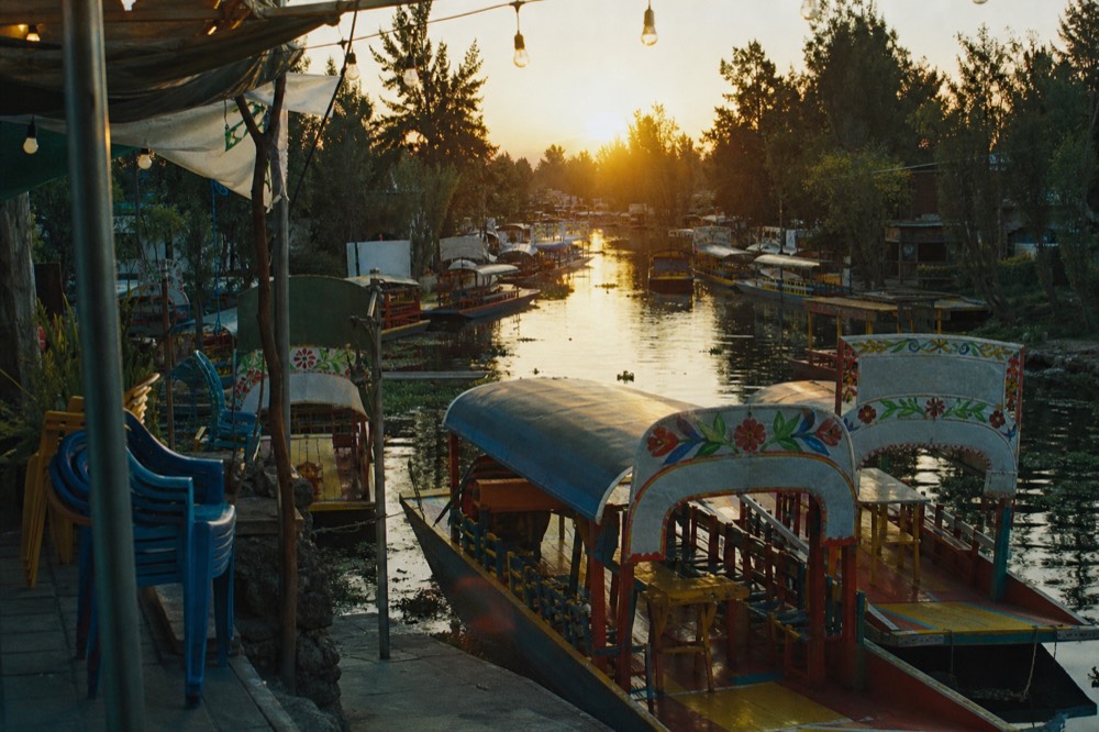 Evening dock with boats moored under strings of bare bulbs, empty plastic chairs stacked at the edge of the landing