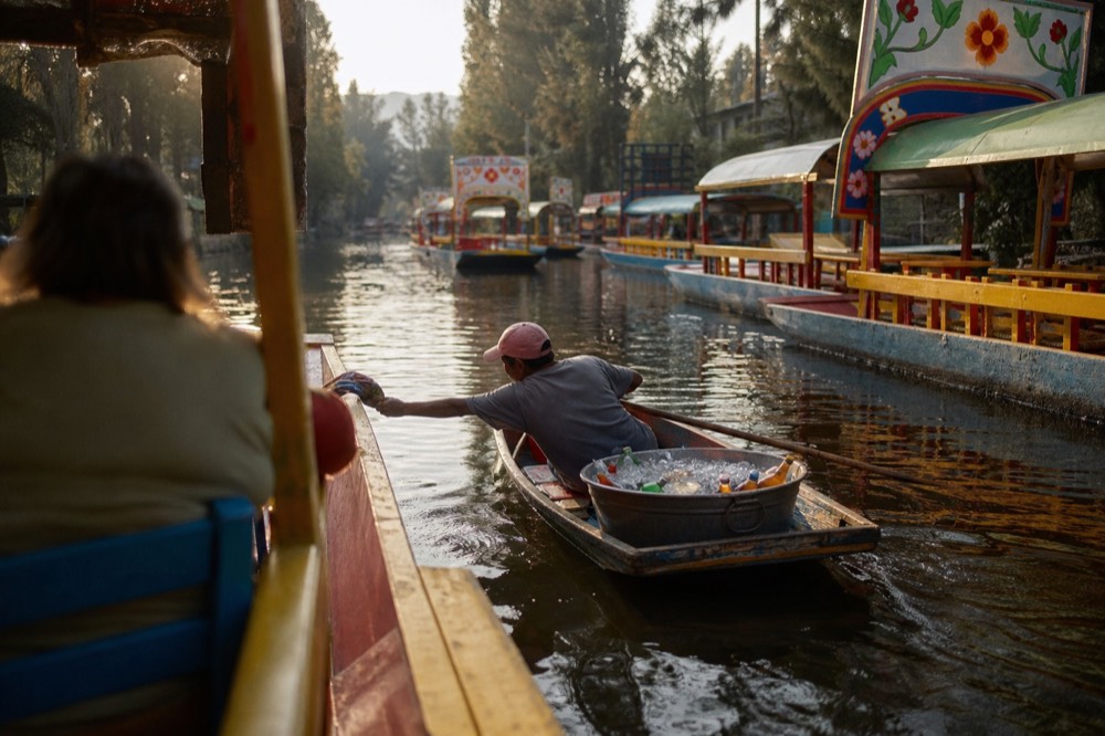 Vendor canoe loaded with a metal tub of iced bottles pulling alongside a larger boat, the paddler leaning across to hand something over