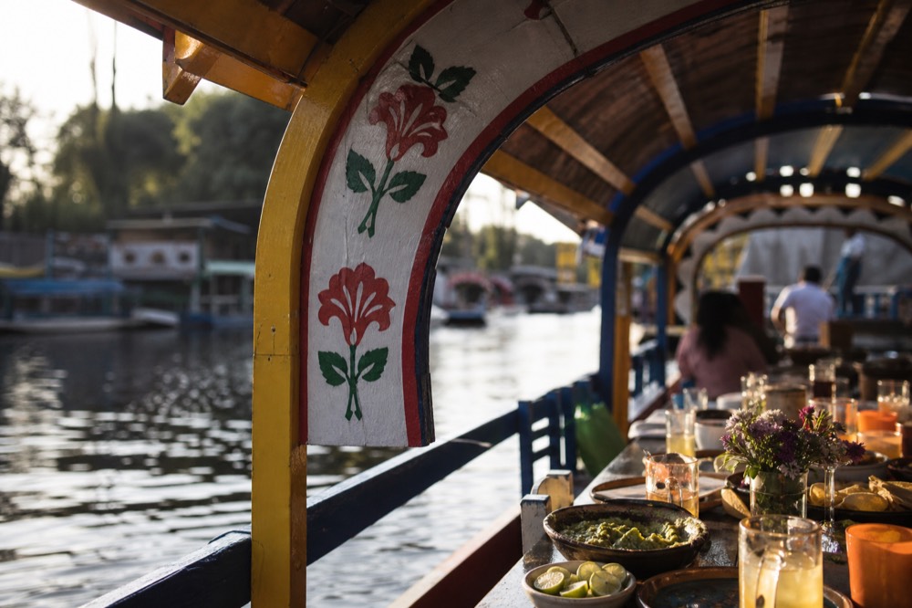 Long communal table on a canal boat set with bowls of guacamole, lime wedges and mixed drinks, the canal water blurred beyond the open side