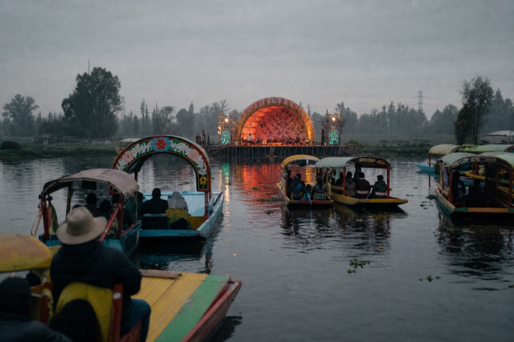 Floating open-air stage on dark water lit by torches and coloured spotlights, silhouettes of boats gathered in a half circle to watch