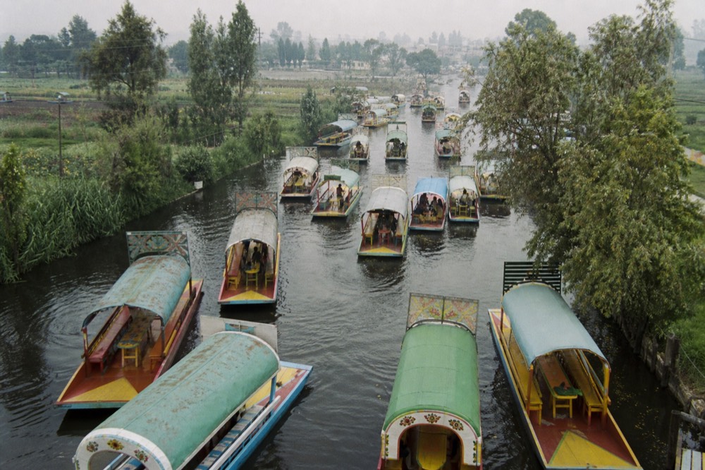 Aerial view of dozens of canopied boats packed nose to tail along a wide canal on a busy afternoon, wakes crossing between them